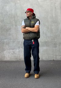 Gilet imbottito verde oliva sopra una camicia bianca, jeans blu scuro, stivali da lavoro color cammello, berretto rosso e occhiali da sole. Accessorio portachiavi visibile.