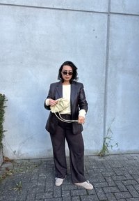 Dark brown leather blazer over a light cream top. Wide brown pants, holding a light beige drawstring bag, wearing black sunglasses.