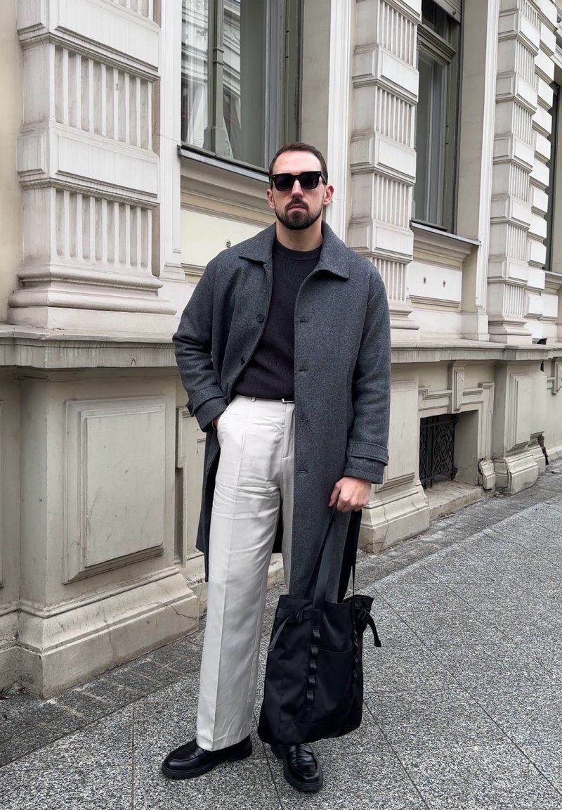 Homme en manteau gris, pull noir, pantalon blanc et chaussures noires, debout sur le trottoir en ville, tenant un sac fourre-tout noir.