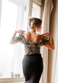 Top de ombro à mostra com estampado de leopardo e mangas com babados, combinado com uma saia preta justa. Janela transparente e plantas de interior ao fundo.