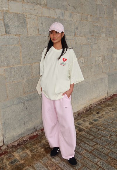 Mujer con camiseta blanca de gran tamaño con diseño de corazón rojo, pantalones de pierna ancha rosa, gorra rosa y zapatillas negras, de pie junto a un muro de piedra.