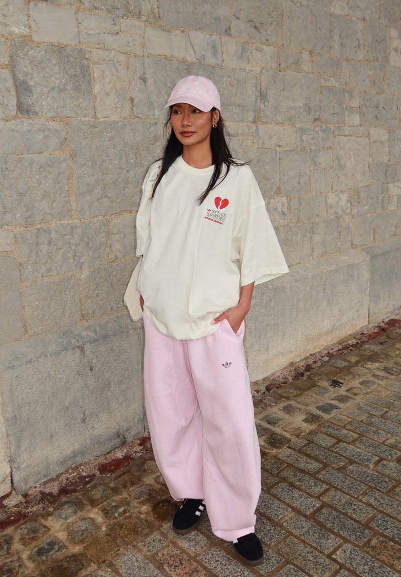 Femme portant un t-shirt blanc ample avec un motif de cœur rouge, un pantalon large rose, une casquette rose et des baskets noires, debout près d’un mur en pierre.