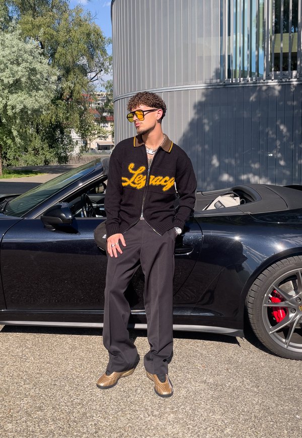 Cardigan preto com fecho e com texto amarelo "Legacy", detalhes em dourado, calças pretas e ténis metálicos castanhos. Carro conversível estacionado ao fundo.