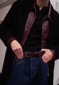 Veste en cuir bordeaux portée sur un t-shirt côtelé noir, associée à un jean taille haute bleu. Bague argentée et ceinture avec une boucle rectangulaire visibles.