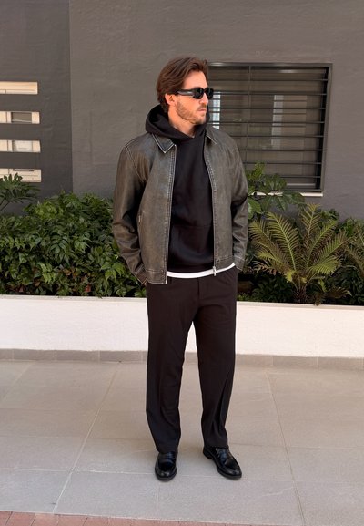 Hombre con gafas de sol negras, chaqueta de cuero negra, sudadera con capucha, pantalones y zapatos negros, de pie al aire libre cerca de plantas y una ventana.