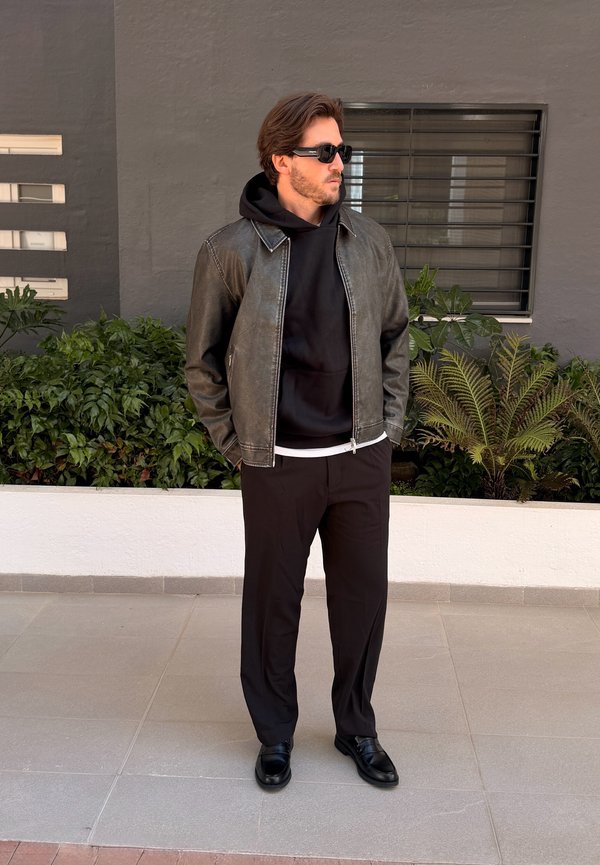 Vyras dėvintis juodus saulės akinius, juodą odinę striukę, džemperį su kapišonu, juodas kelnes ir batus, stovintis lauke šalia augalų ir lango.