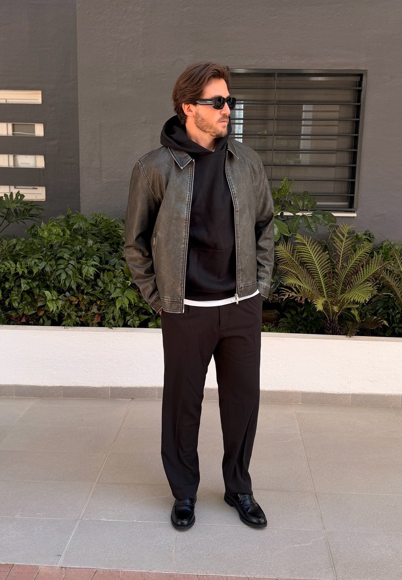 Homme portant des lunettes de soleil noires, une veste en cuir noire, un sweat à capuche, un pantalon et des chaussures noirs, debout à l'extérieur près de plantes et d'une fenêtre.