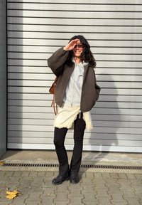 Femme aux longs cheveux bouclés, portant des lunettes de soleil, une veste à carreaux, un sweat à capuche gris, une jupe crème sur un pantalon noir, debout sur un trottoir près d'un mur gris.