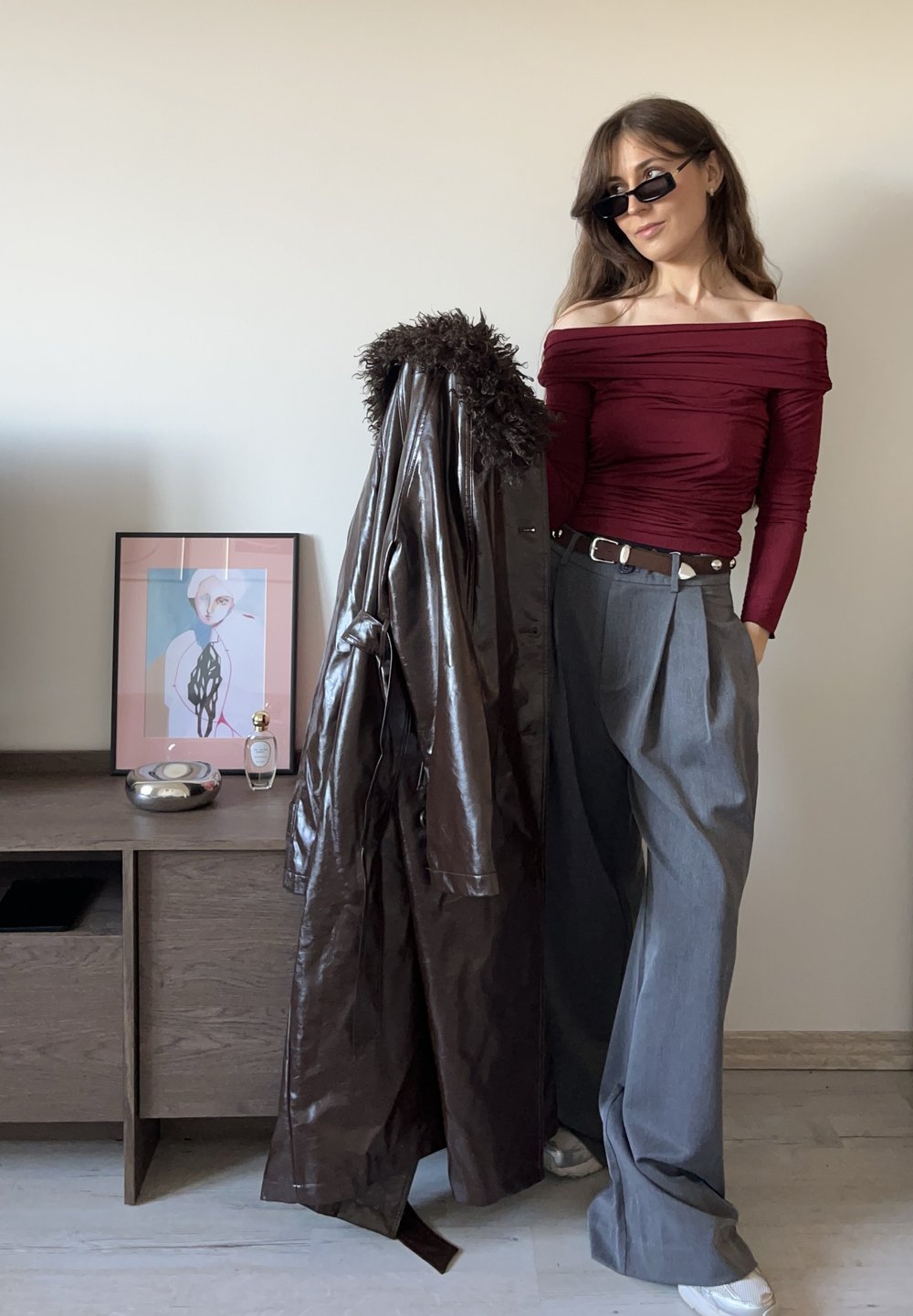 Cappotto marrone in ecopelle con dettagli in pelliccia, top bordeaux scivolato sulle spalle e pantaloni grigi a pieghe. Occhiali da sole neri completano il look.