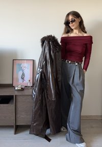 Cappotto marrone in ecopelle con dettagli in pelliccia, top bordeaux scivolato sulle spalle e pantaloni grigi a pieghe. Occhiali da sole neri completano il look.