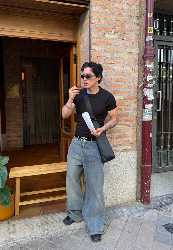 Persona con gafas de sol y camisa negra está de pie frente a un edificio de ladrillo comiendo un alimento, sosteniendo una bolsa de papel y llevando una bolsa negra al hombro.