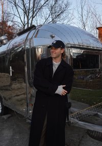 Un rimorchio Airstream vintage d'argento, forma arrotondata, esterno lucido, con superfici riflettenti, posto davanti a uno sfondo di rami d'albero.