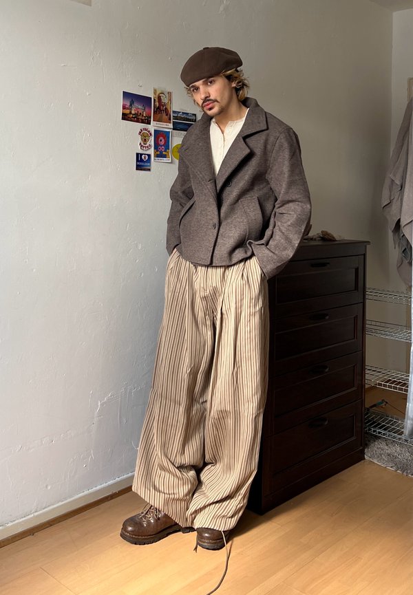 Joven con boina marrón, chaqueta gris, pantalones anchos beige con rayas y botas marrones, de pie con las manos en los bolsillos cerca de una cómoda.