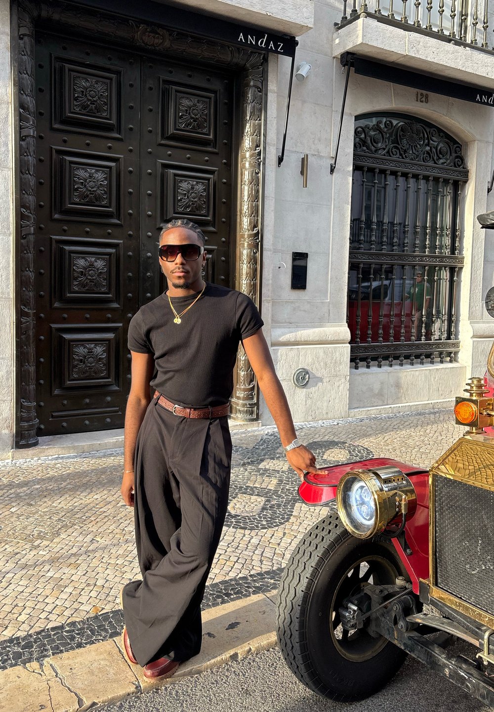 Maglietta nera a coste, pantaloni neri larghi, cintura marrone. Auto d'epoca rossa con dettagli dorati e proiettore rotondo parcheggiata accanto a una porta nera ornata.