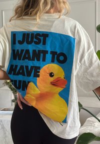 Camiseta oversized branca com um fundo azul, apresentando um gráfico de um pato de borracha amarelo e texto em negrito preto que diz "EU SÓ QUERO ME DIVERTIR."