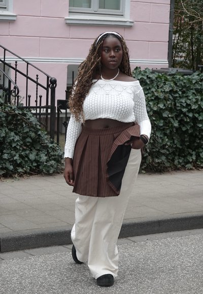 Jersey corto blanco de punto con mangas largas, falda marrón con pliegues y rayas, pantalones blancos holgados y zapatos negros; de pie al aire libre.
