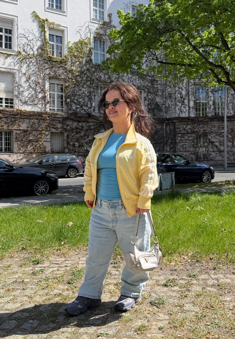 Mujer con cabello rizado que lleva gafas de sol, chaqueta amarilla, camisa azul, vaqueros claros y zapatillas, de pie sobre la hierba cerca de un edificio y coches aparcados.