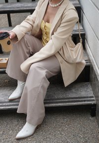 Bež blazer, žuta bluzica s gumbima, bež široke hlače, bijele ukazne čizmice, i bež torbica; drži šalicu kave.