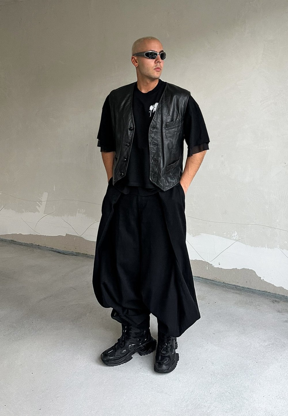 Μαύρο δερμάτινο γιλέκο πάνω από ένα μαύρο t-shirt, συνδυασμένο με φαρδιές μαύρες παντελόνες και χοντρά μαύρα μποτάκια. Λείες υφές και μινιμαλιστικός σχεδιασμός.