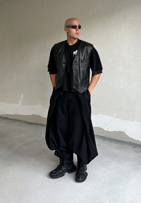 Μαύρο δερμάτινο γιλέκο πάνω από ένα μαύρο t-shirt, συνδυασμένο με φαρδιές μαύρες παντελόνες και χοντρά μαύρα μποτάκια. Λείες υφές και μινιμαλιστικός σχεδιασμός.