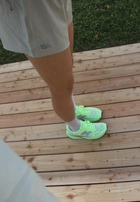 Neon grønne sportssko med et mesh-design, reflekterende detaljer og hvide sokker. Person iført lyse shorts står på træterrasse.
