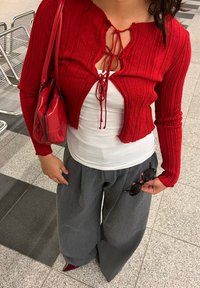 Rød ribbet strikket cropped cardigan med båndlukning, båret over en hvid bluse. Sæt sammen med grå vidde bukser og holder solbriller.