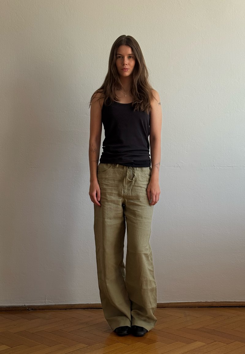Jeune femme aux longs cheveux bruns se tient devant un mur uni, portant un haut noir sans manches, un pantalon beige ample et des chaussures noires sur un sol en bois.