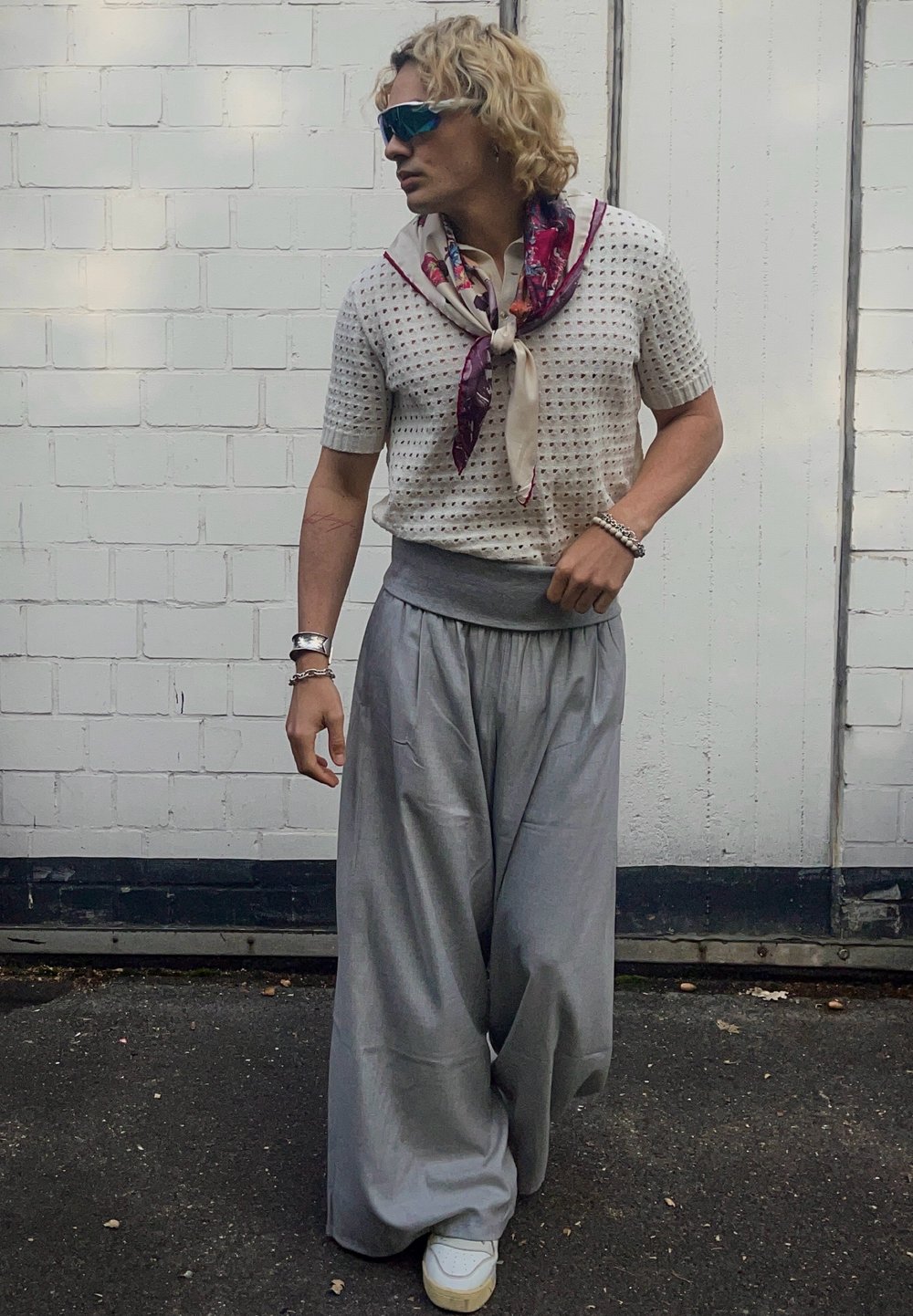 Világosszürke, bőszárú nadrág magas derékkal, amelyhez egy rövid, fehér perforált kötött felsőt és nyaknál megkötött mintás sálat visel.