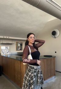 Διάφανο καφέ μπλούζα μακρύ μανίκι με διακοσμητικά μοτίβα πάνω από ένα μαύρο εφαρμοστό μπλουζάκι, συνδυασμένο με ανοιχτό γκρι τζιν και μια λευκή τσάντα ώμου.