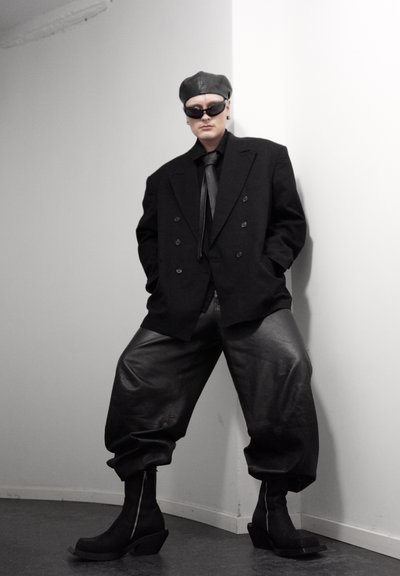 Persona con blazer negro de gran tamaño, pantalones de cuero, botas negras, gafas de sol y gorra de cuero, de pie contra una pared blanca con esquina.