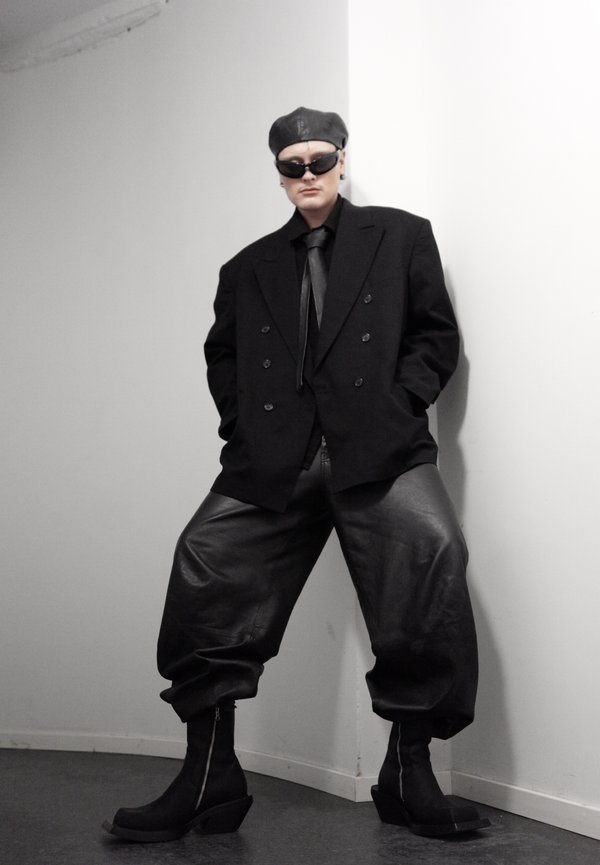Person iført oversized sort blazer, læderbukser, sorte støvler, solbriller og læderkasket, stående mod en hvid hjørnevæg.