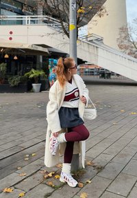 Palton alb lung peste un pulover vișiniu cu dungi, fustă în dungi, colanți vișinii și adidași albi cu accente roșii. Accesoriu: geantă albă.