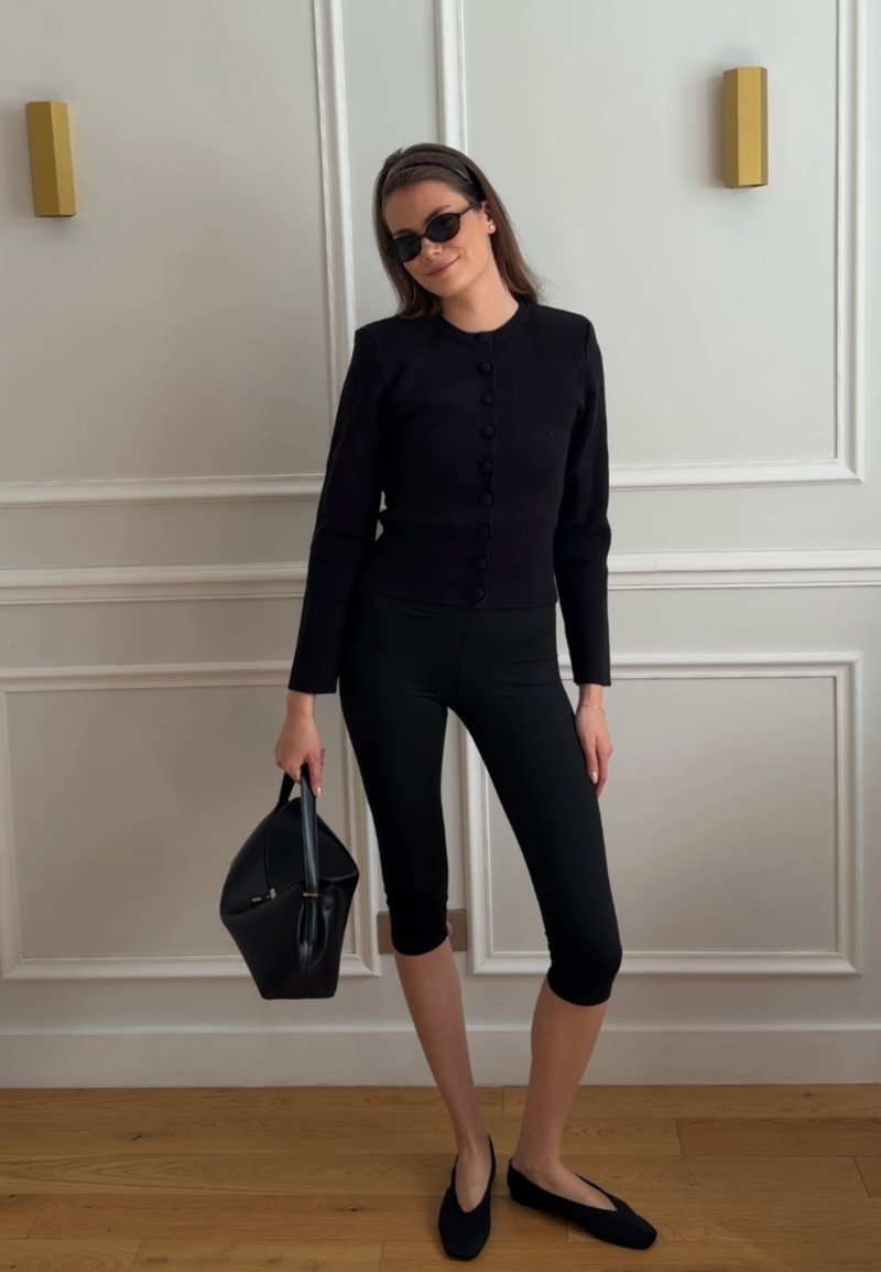 Femme portant une veste noire boutonnée, des leggings courts, des ballerines noires, des lunettes de soleil, tenant un sac à main noir, debout contre un mur blanc à panneaux.