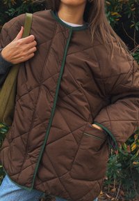 Barna steppelt dzseki zöld díszítéssel, laza szabással és oldalsó zsebekkel, szürke pulóver fölött viselve, kültéri környezetben bemutatva.