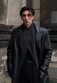 Svart skinnkåpe over en mørk tekstilantrekk, med sjalkrage og knappedetaljer. Glatt tekstur og tettsittende design.