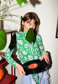 Vestido verde de malha com padrões florais, segurando um disco de vinil preto com um rótulo vermelho. O fundo inclui uma planta e uma prateleira vermelha.