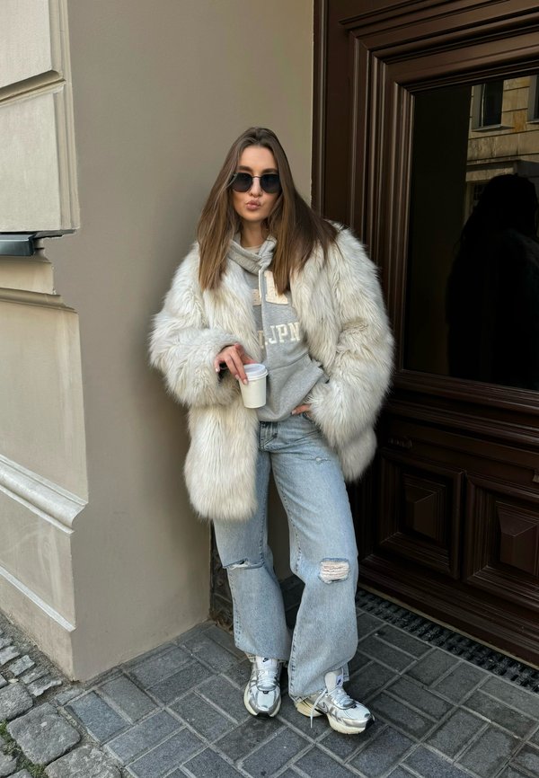 Abrigo de piel sintética blanca, sudadera gris, jeans claros rasgados y zapatillas plateadas. De pie contra una pared marrón, sosteniendo una taza blanca.