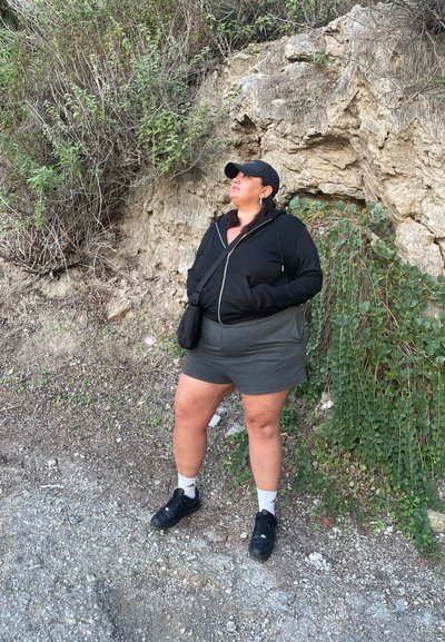 Sudadera negra con cremallera, pantalones cortos grises, zapatillas negras y calcetines blancos. Entorno natural con un fondo rocoso y vegetación verde.