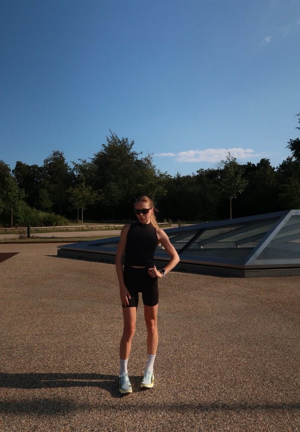 Jeune personne en tenue de sport noire et lunettes de soleil se tenant à l'extérieur sur une surface pavée avec des arbres et une structure en verre en arrière-plan.