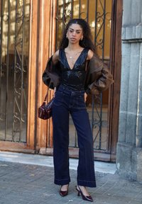 Schwarzes Top mit Pailletten, braune übergroße Jacke, dunkelblaue Schlagjeans, kastanienbraune Handtasche und bordeauxrote High Heels. Städtische Umgebung mit Holztüren.