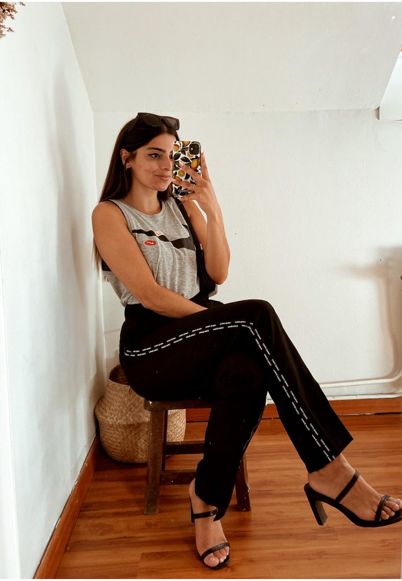 Szürke ujjatlan felső piros akcentussal, fekete nadrág fehér oldalsávokkal, és fekete magassarkú szandál. Fából készült széken ül.