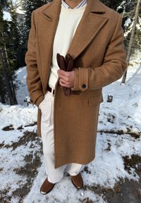 Άνδρας που φοράει καφέ παλτό, λευκό πουλόβερ, λευκό παντελόνι και καφέ παπούτσια, στέκεται σε χιονισμένο έδαφος κρατώντας καφέ δερμάτινα γάντια.