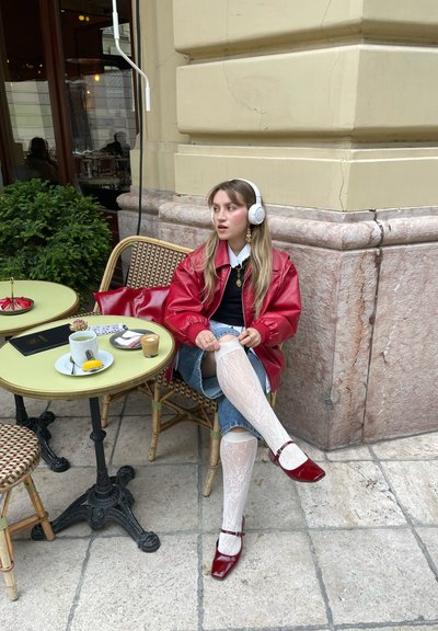 Chaqueta bomber roja, suéter negro, falda de mezclilla. Medias de rayas blancas y zapatos rojos de punta cuadrada. Mesa redonda con café y postre.