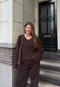 Maglione oversize bordeaux con texture pelosa e maniche larghe, abbinato a pantaloni marrone scuro. Borsa intrecciata nera, in piedi accanto a una porta.