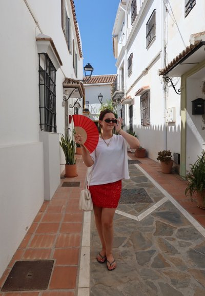 Abanico plegable rojo, blusa corta blanca de mangas cortas y falda roja con lentejuelas. Paseo pavimentado, edificios blancos con barandillas negras y plantas verdes.