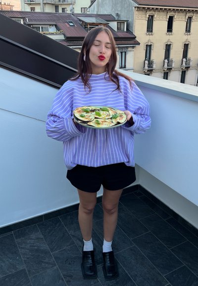 Suéter oversized a rayas moradas y blancas, pantalones cortos negros, sosteniendo un plato de comida verde y blanca en una azotea con un fondo urbano.