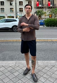 Braun gestreiftes Langarmhemd, dunkelblau Shorts, schwarze Slipper und sichtbares Beintattoo; steht auf einem Bürgersteig in der Stadt.