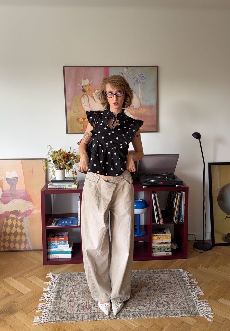 Persona con pantaloni beige oversize e camicetta nera a pois che si trova in una stanza con quadri incorniciati e un giradischi su una mensola.