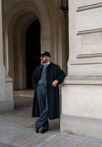 Cappotto nero lungo sopra un maglione grigio, pantaloni grigi e scarpe nere. Cappello indossato, appoggiato a una colonna di pietra in un contesto urbano.