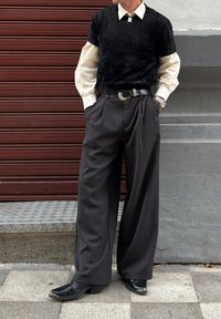 Persoon draagt zwart textuur kortermig shirt over een wit shirt met lange mouwen, wijde grijze broek, zwarte laarzen, zilveren horloge en riem, staand buiten.